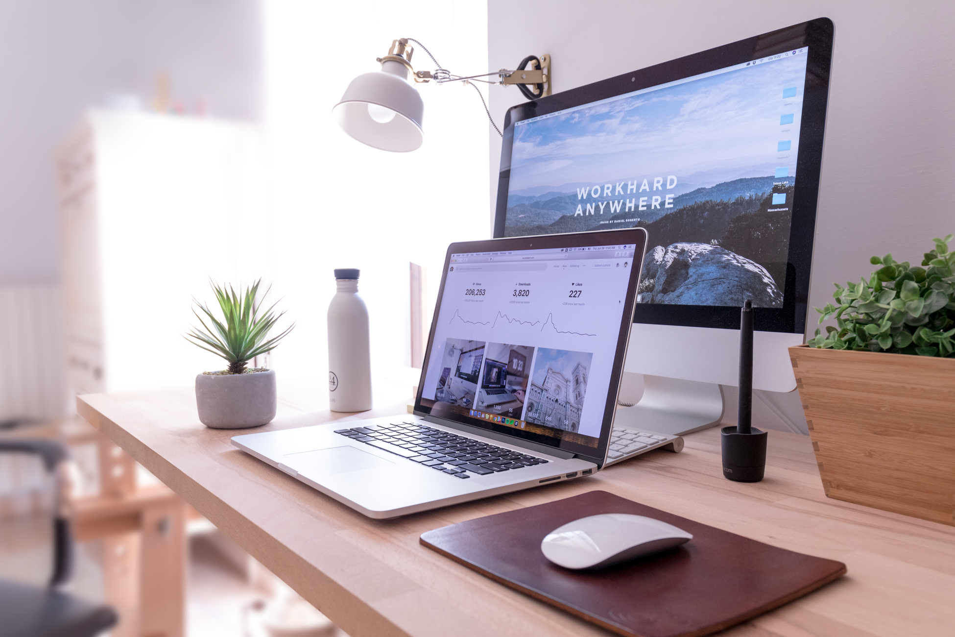 A laptop and desktop computer sit on a minimalist desk with light shining in from a window.